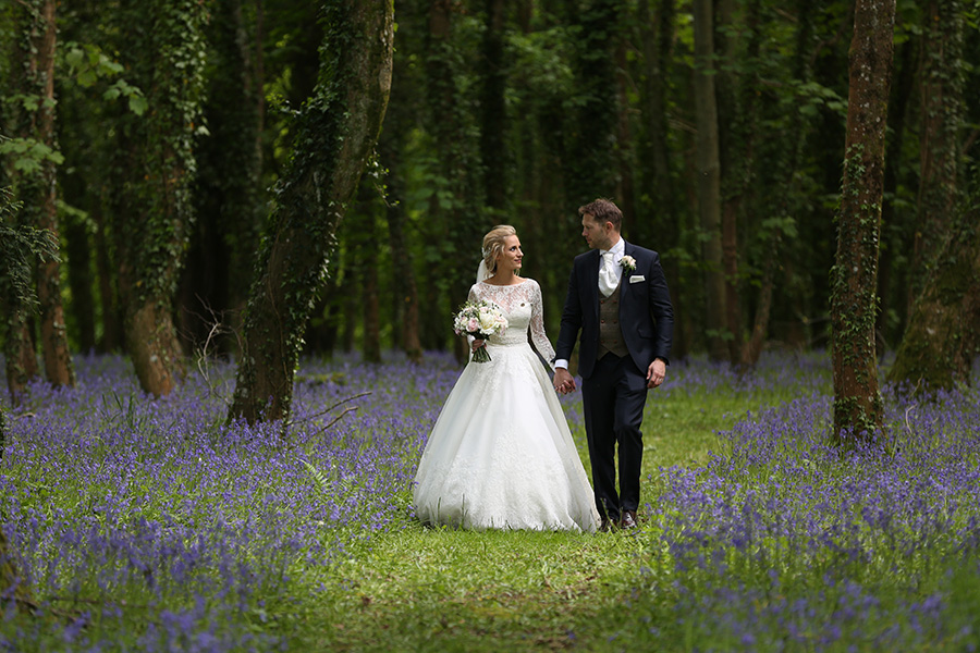 Bride and groom walking hand in hand through woodland bluebell setting during romantic Hampshire wedding.