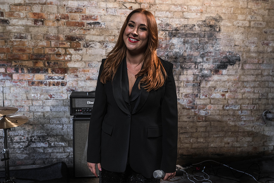 Lyndsey Cooper standing with a microphone in a black outfit against a rustic brick wall, ready to perform at a wedding or live event