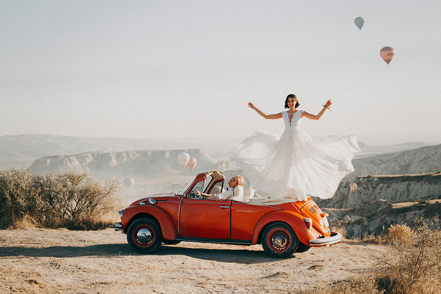 Bride celebrating in flowing wedding dress standing on vintage convertible car overlooking scenic Hampshire countryside landscape.