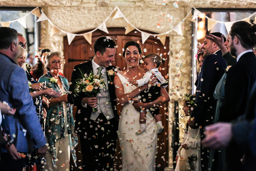 Bournemouth wedding photographer capturing bride and groom with baby walking through confetti at wedding ceremony