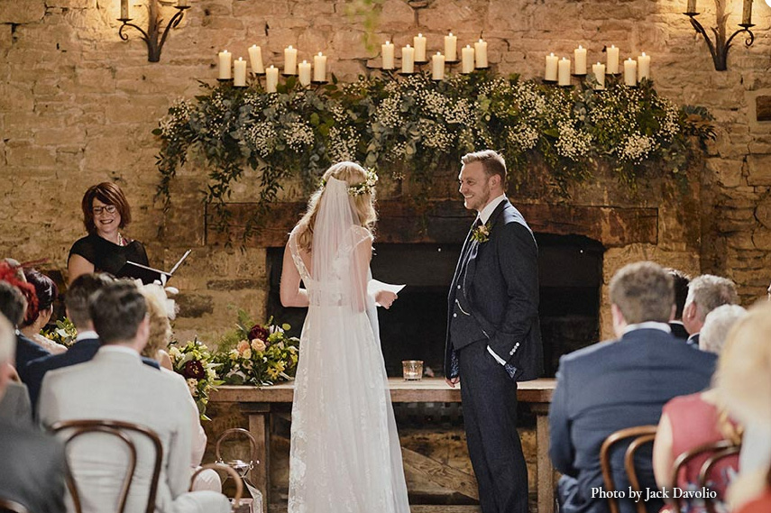 Indoor wedding ceremony with celebrant leading couple in rustic venue with candles and floral fireplace