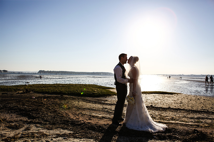 Bride and groom standing together on a beach at sunset with backlit sunlight