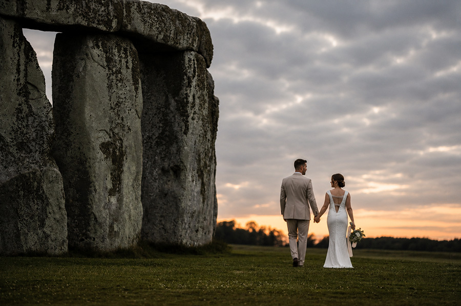Bride and groom walking hand in hand beside Stonehenge at sunset in Wiltshire